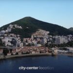 Aerial view of a coastal city with a mountainous backdrop. The image highlights a 'MONTECO' location marker in the city center. The foreground features historic buildings and a marina, with modern buildings and lush green hills in the background.