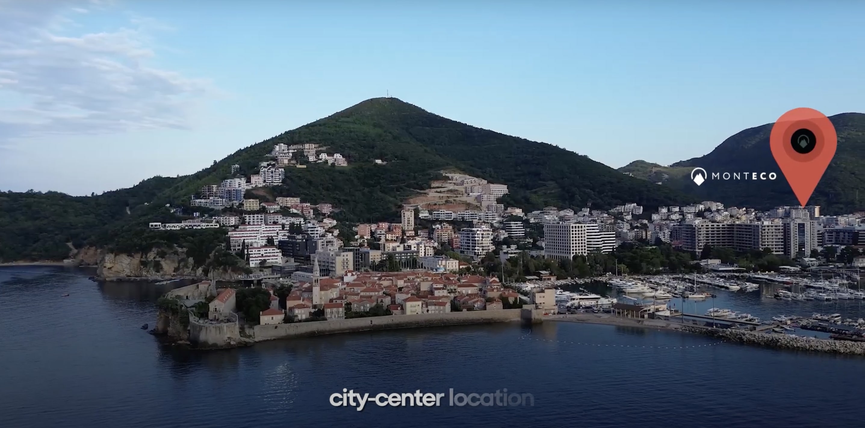 Aerial view of a coastal city with a mountainous backdrop. The image highlights a 'MONTECO' location marker in the city center. The foreground features historic buildings and a marina, with modern buildings and lush green hills in the background.