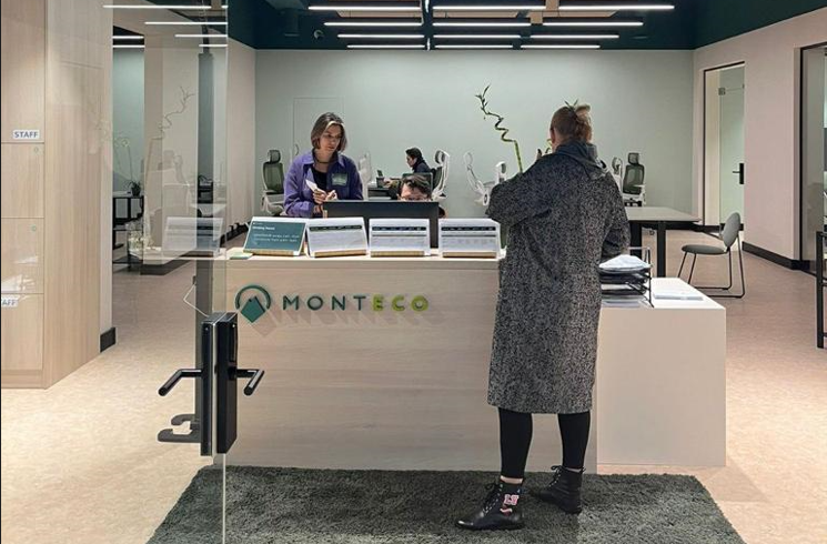 Reception area of a modern co-working space labeled 'MONTECO.' A receptionist is assisting a visitor across a counter. The space features a clean design with light-colored walls and furniture. In the background, people are working at desks with ergonomic chairs, creating a professional and welcoming environment.