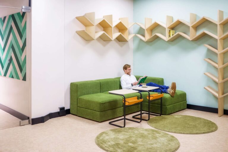 Man relaxing on a green couch in a modern co-working space. The area features stylish geometric wall shelves, circular green rugs, and contemporary decor with a green and white patterned wall. The man is using a tablet and appears comfortable, creating a relaxed and productive atmosphere.
