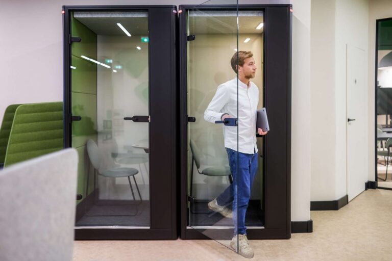 Man exiting a private phone booth in a modern co-working space. The booth is equipped with a chair and a small table, designed for quiet and focused work or calls. The man is holding a laptop and wearing casual attire, walking into the main area of the co-working space, which features stylish green seating.