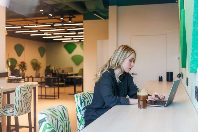 Woman working on a laptop at a counter in a modern co-working space. She has a drink next to her and is focused on her work. The background features stylish decor with green accents, plants, and well-lit workstations, creating a productive and comfortable environment.