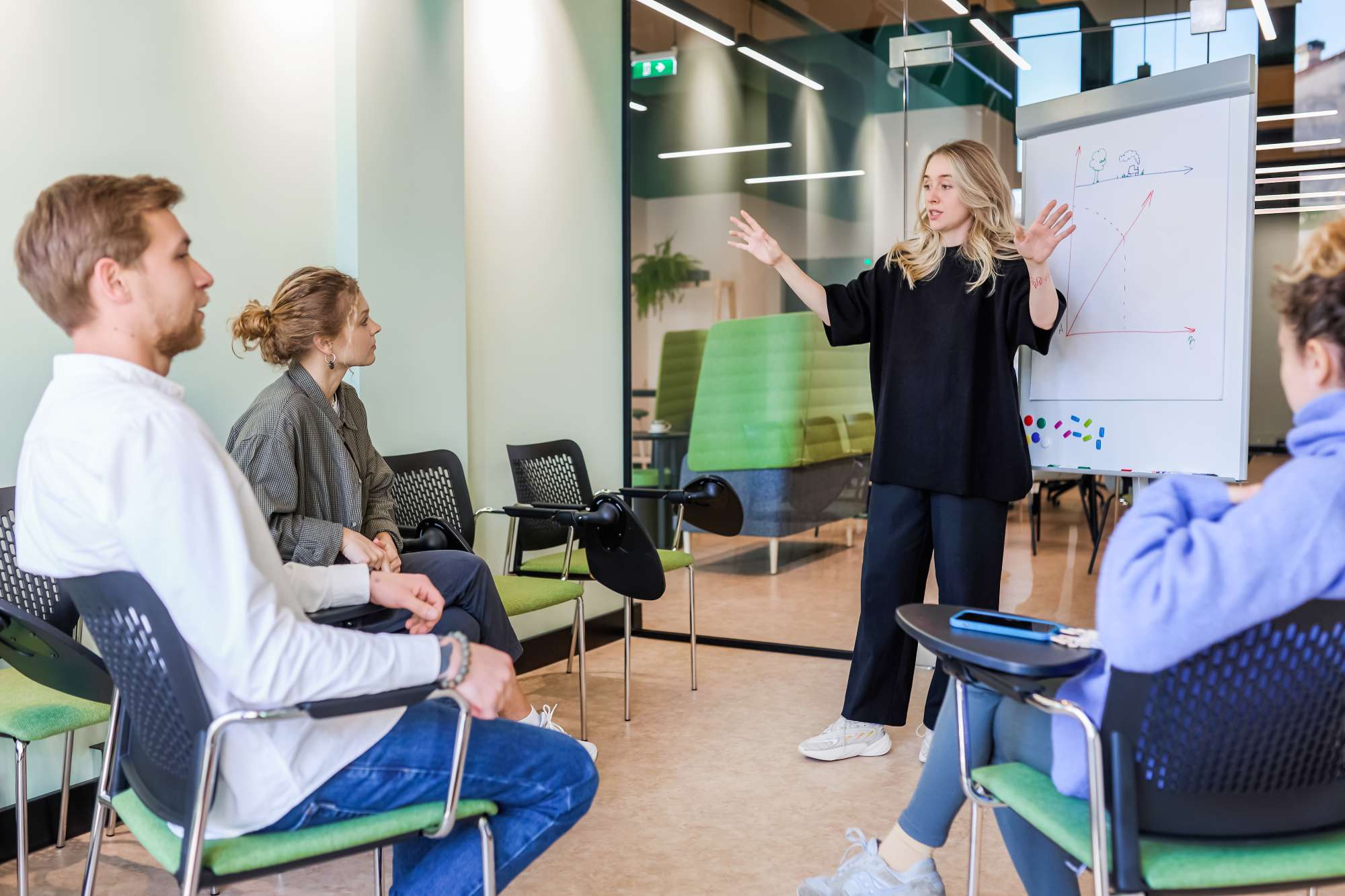 Predavaonica Woman giving a presentation to a small group in a modern co-working space. The presenter is standing next to a whiteboard with diagrams, engaging the audience with her gestures. The attendees are seated in a semi-circle, listening attentively. The room has a bright, open design with glass partitions and green accents.