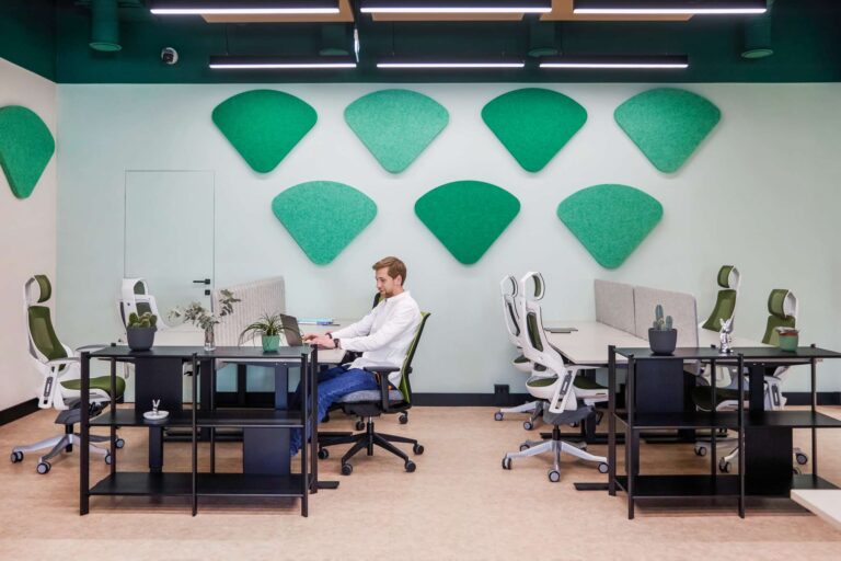 Man working on a laptop in a spacious, modern co-working space. The room features green geometric wall decorations, ergonomic office chairs, and individual workstations with plants adding a touch of nature. The clean and well-organized environment provides a productive and comfortable atmosphere.