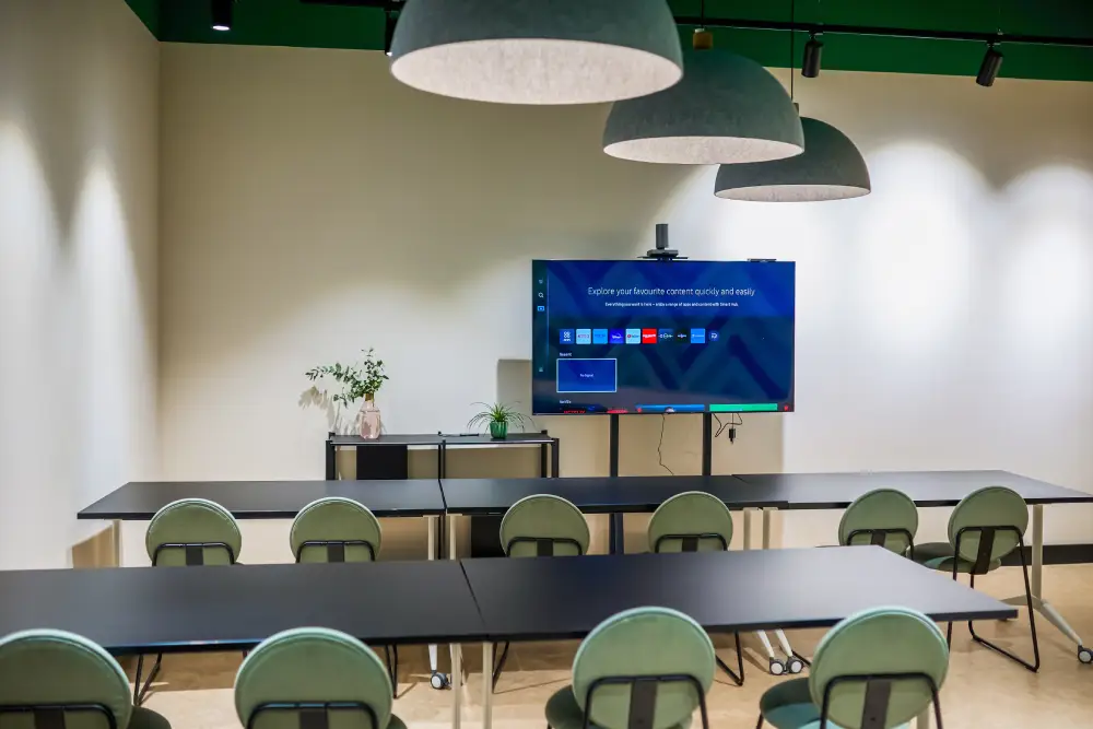MONTEHUB Empty modern co-working space set up for a presentation or meeting. The room features a large screen displaying a menu, several tables with green chairs, and stylish pendant lights. A small table with plants adds a touch of greenery, creating a professional yet inviting atmosphere.