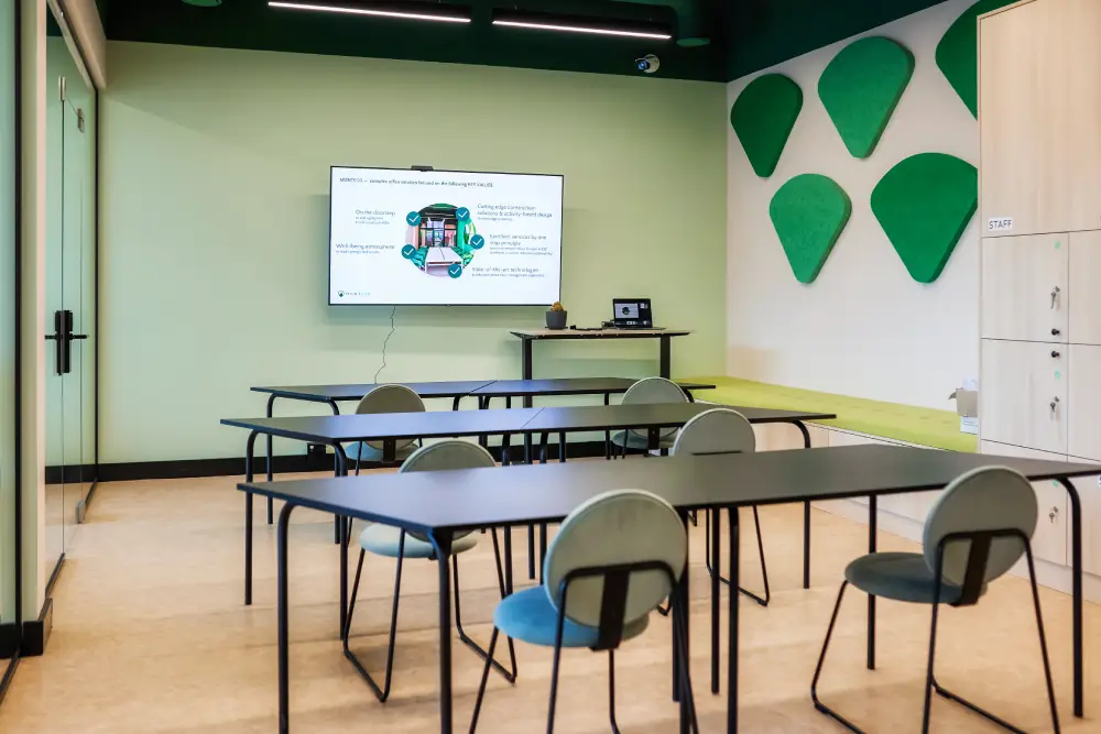 MONTEHUB Empty modern co-working space with a presentation setup. The room features a large screen displaying a presentation, several tables with chairs, and green geometric wall decorations. The space is well-lit and organized, with lockers labeled 'Staff' on the right and a minimalistic design that fosters productivity.
