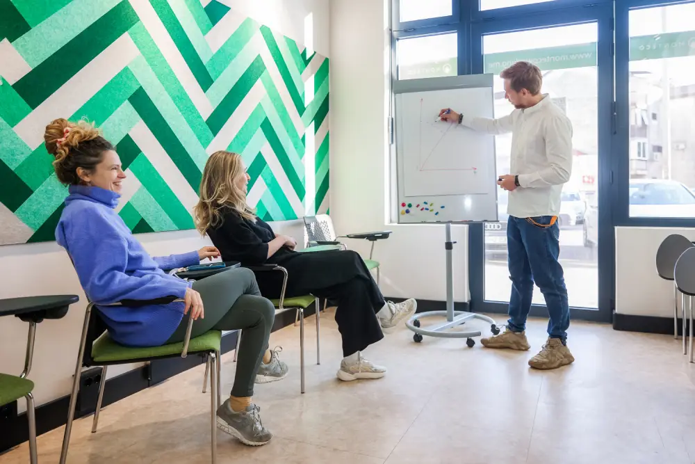 MONTEHUB Man giving a presentation to two women in a modern co-working space. The presenter is writing on a whiteboard, while the women sit on green chairs and listen attentively. The room features a large window and a vibrant green and white patterned wall, creating a bright and engaging environment.